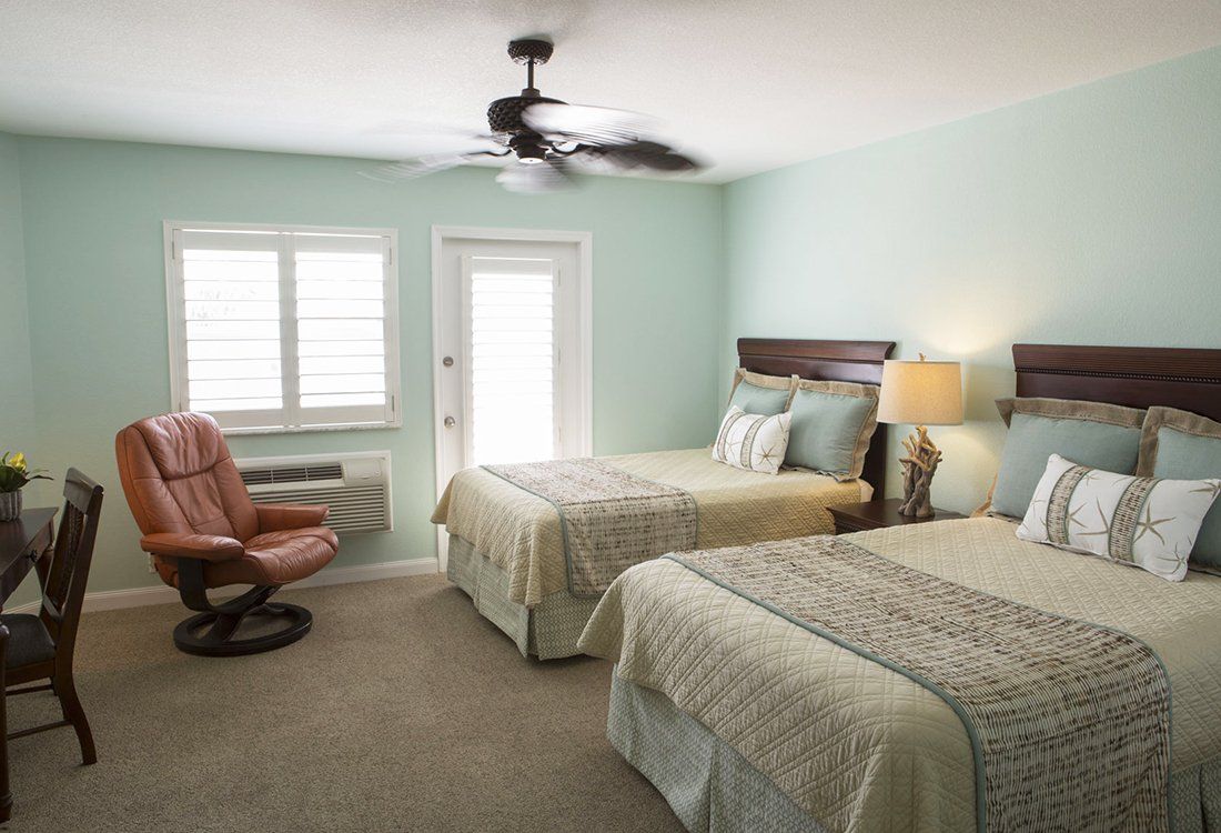 A bedroom with two beds, a recliner, and a door to a balcony. Light blue walls, beige bedding, and brown furniture.
