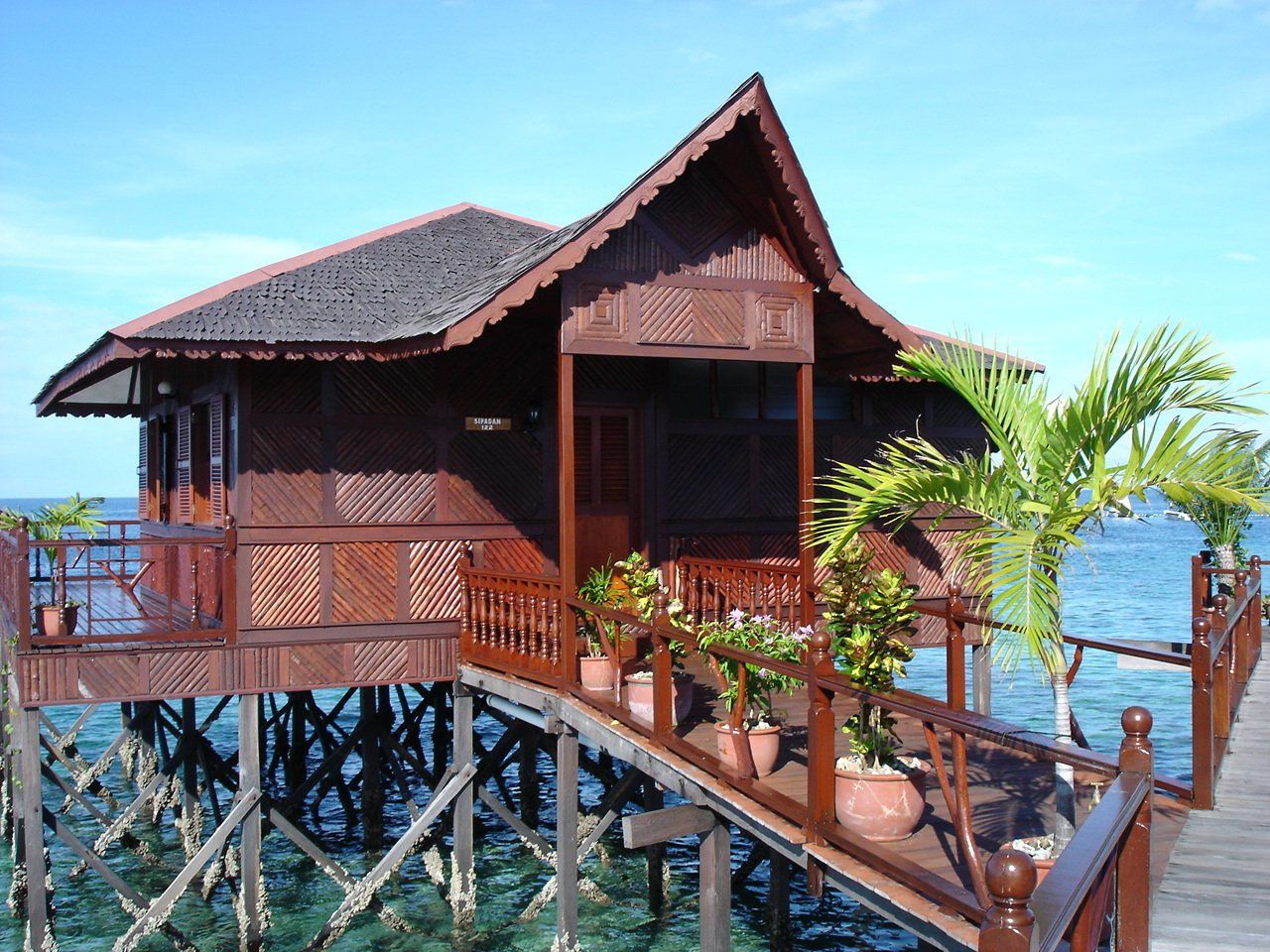Wooden overwater bungalow with brown roof and railings, on stilts above turquoise water.