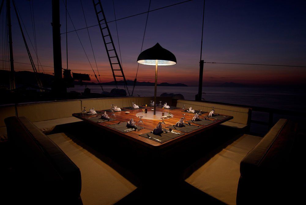 Outdoor dining table set for a meal on a boat deck at dusk, with the sea and sky in the background.
