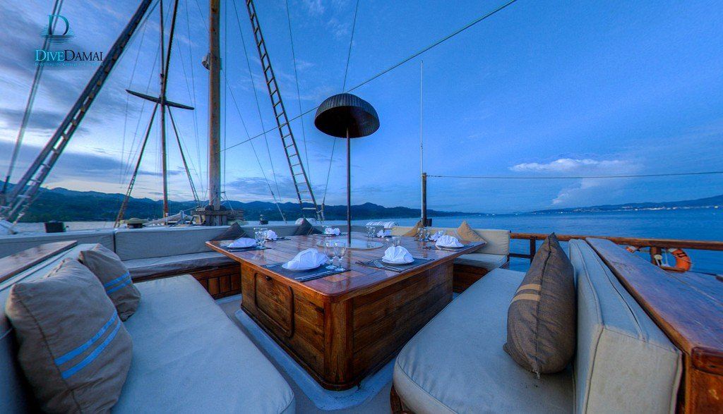Outdoor dining area on a sailboat at dusk, with table, cushions, and distant water.