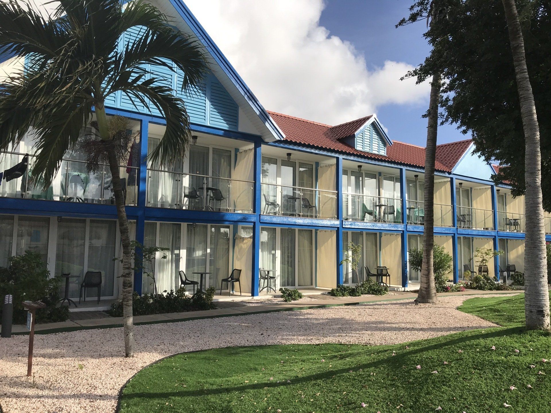 Two-story hotel building with blue accents, white walls, and red tiled roof, surrounded by palm trees and landscaping.
