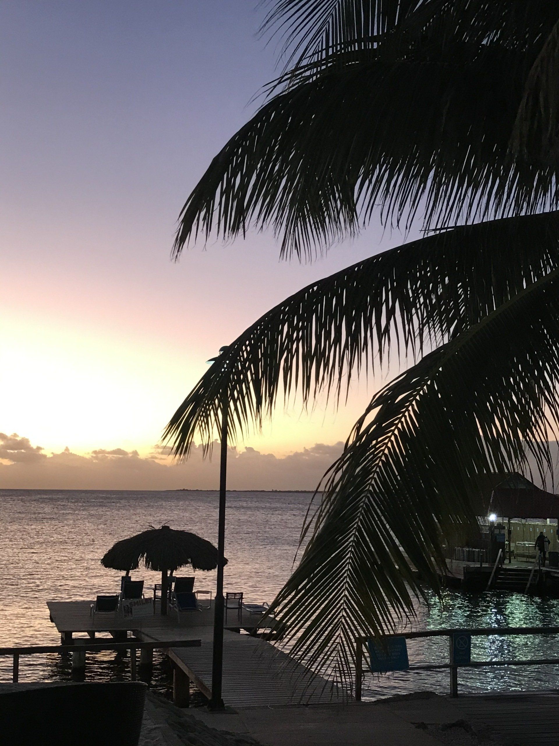 Palm trees frame a serene sunset over the ocean. Pier with a palapa and chairs in silhouette.
