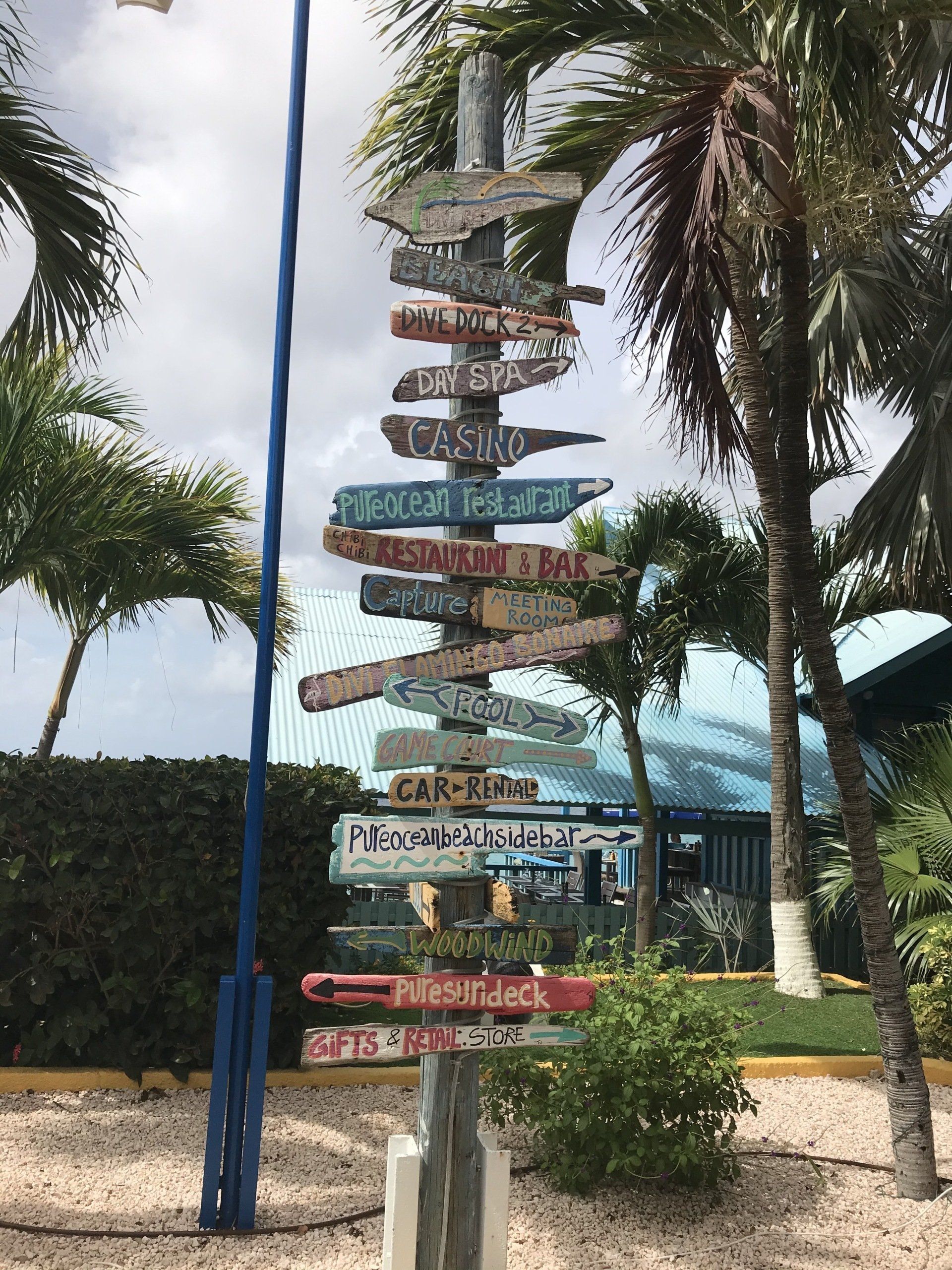 Tall post with many signs pointing in different directions. Outdoor setting with palm trees and a building.