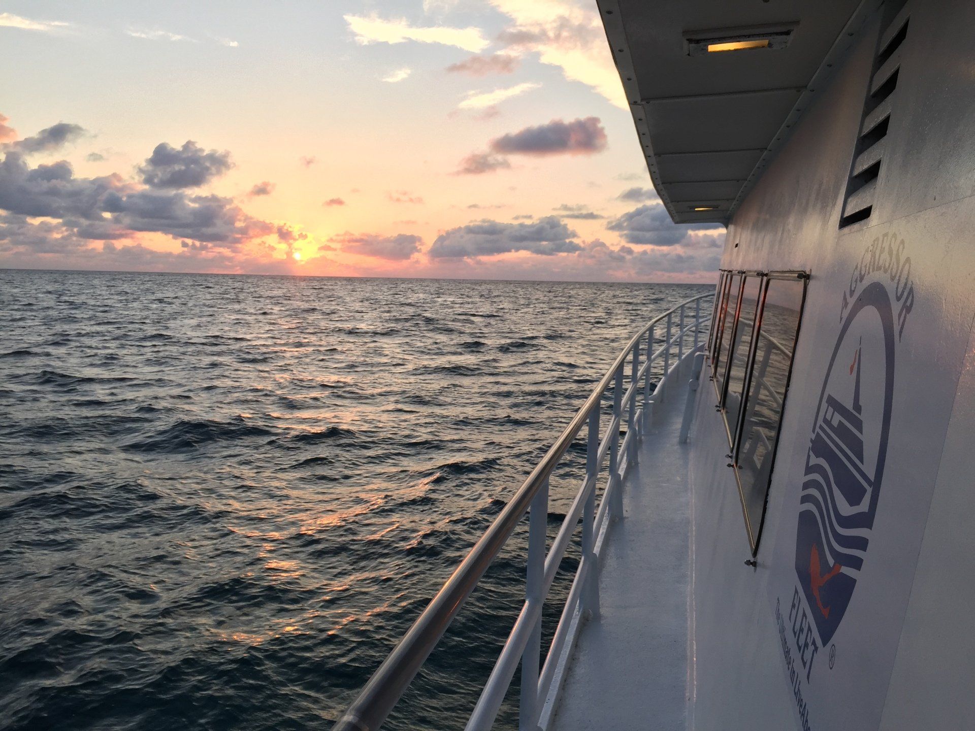 Sunset over the ocean, viewed from the side of a white boat with a railing. Clouds and a logo are visible.