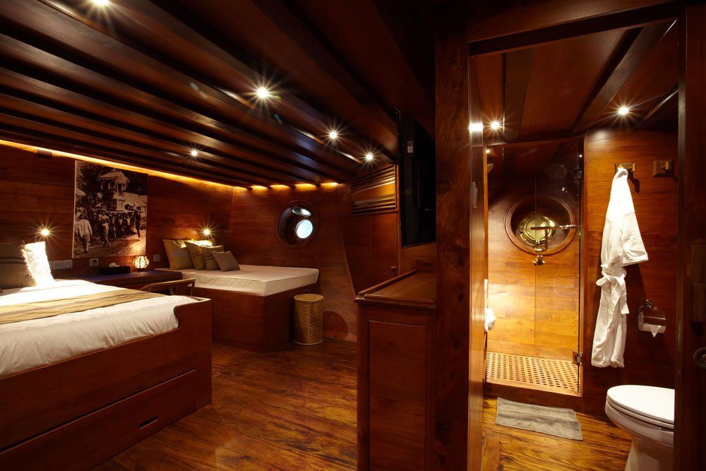 Wooden ship cabin with a bed, porthole, and bathroom. Warm lighting and a robe hanging.