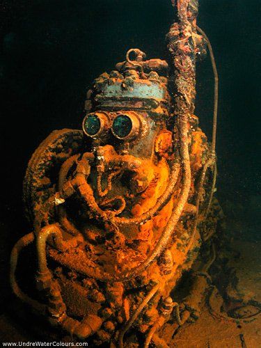 An old, rusty diving helmet and equipment underwater. Covered in marine growth, dark water background.
