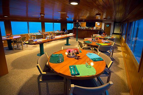 Dining room inside a boat with round and rectangular tables, green placemats, and ocean views.
