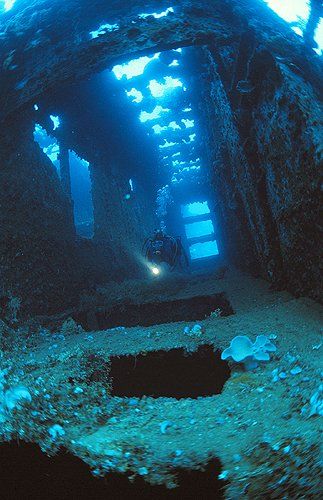 Underwater view of a sunken shipwreck's interior, illuminated by a diver's flashlight. Blue water and open hatches.