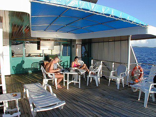 Deck of a boat with people sitting at a table under a blue canopy, with the ocean in the background.