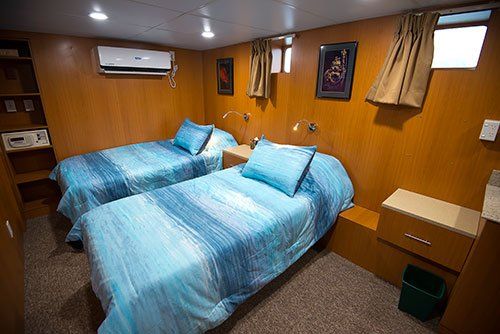 Two twin beds in a wood-paneled room on a boat; blue bedding, air conditioner, small windows.