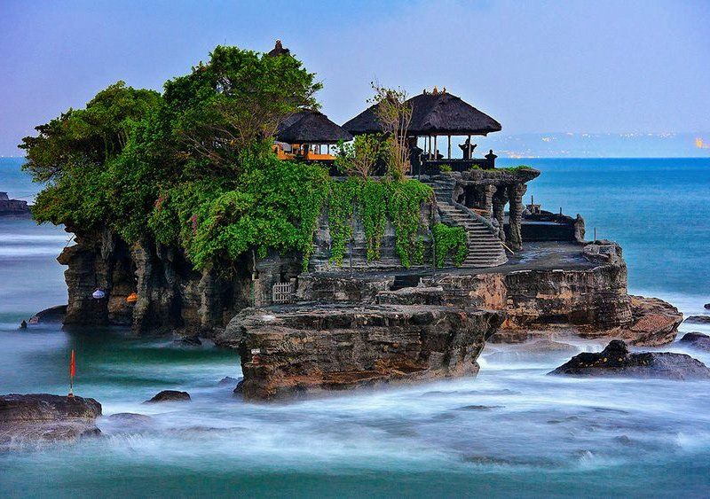 Tanah Lot temple on a rocky island in the ocean, Bali. Stone structure with thatched roofs; waves blur around it.