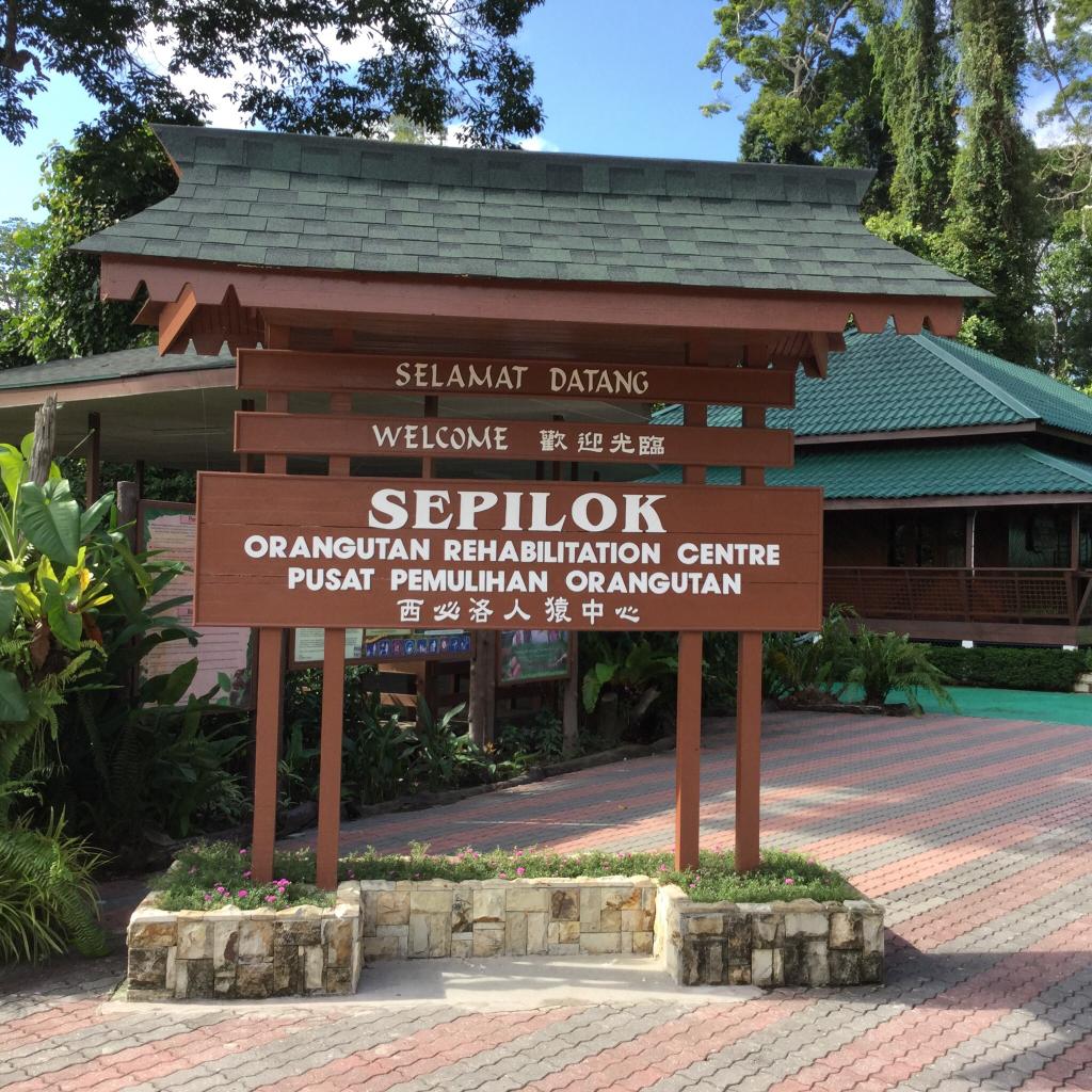 Sign for Sepilok Orangutan Rehabilitation Centre, brown with green roof and text in multiple languages.