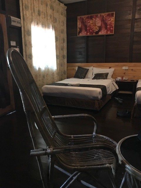 Wicker chair in front of a bedroom with dark wood paneling, a bed, and a window with sheer curtains.