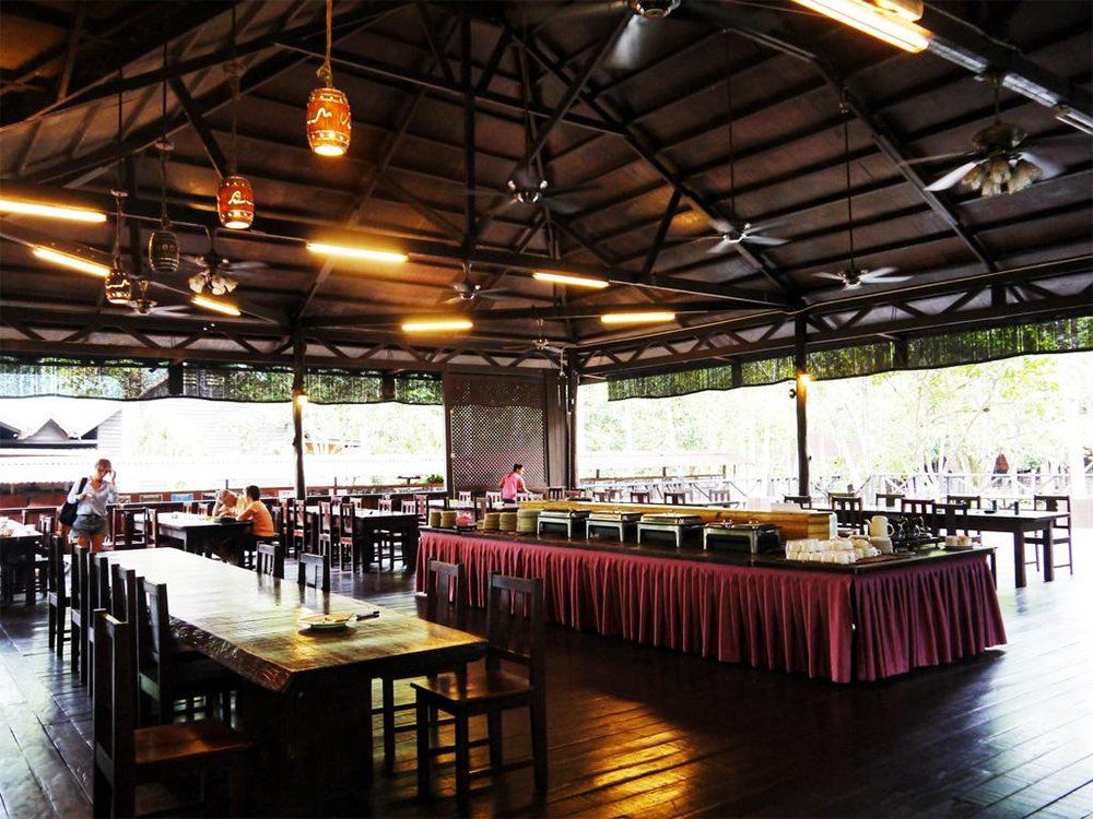 Large, open-air dining hall with tables, buffet, and ceiling fans. Guests seated, eating.