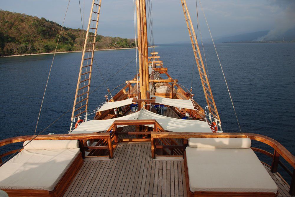 View from the wooden deck of a boat towards the island coast. White cushions, blue water, and brown wood.