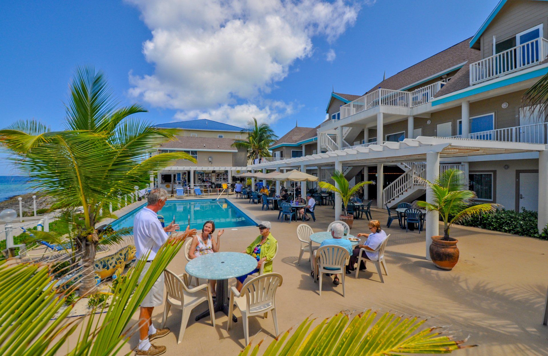 Outdoor patio and pool area; people at tables, man gesturing. Blue sky, buildings.