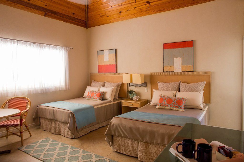 Bedroom with two beds, beige walls, wooden ceiling. Each bed has a blue blanket and colorful pillows.