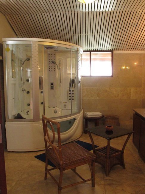 A modern bathroom with a large shower unit, chair, small table, and a window.