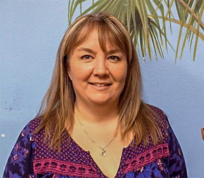 Woman with blonde-streaked hair, smiling, wearing a patterned top, in front of a blue wall with a palm tree.
