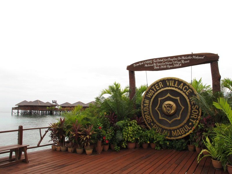 Water village resort entrance with a large gong, overwater bungalows, and lush greenery.