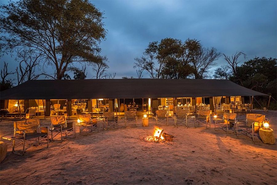 Camp dining area at dusk with a bonfire and chairs; trees in the background.