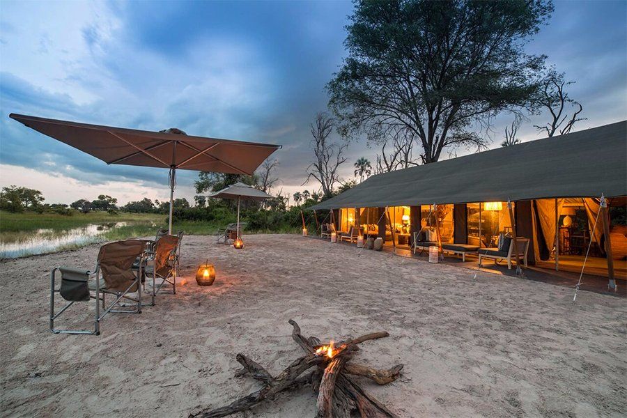 Campsite in Africa at dusk, with bonfire, chairs, tents, and large umbrella over the sand.