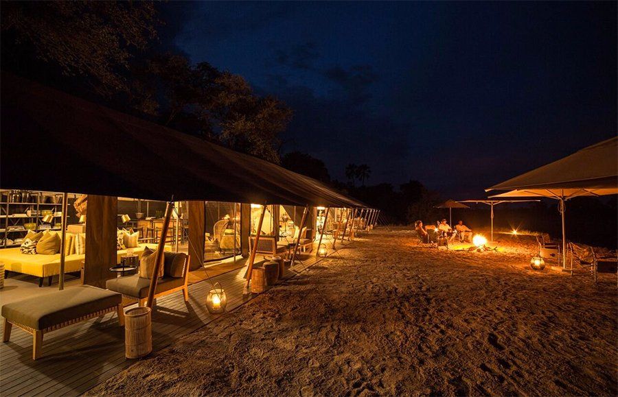Nighttime outdoor dining area with lit tents, a fire pit, and tables.