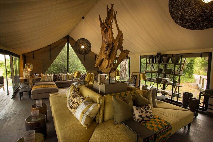 Lounge area inside a tent with wood furniture, yellow sofa, and natural light from large windows.