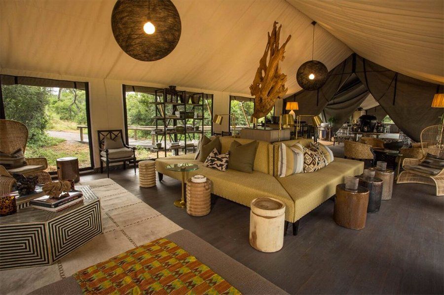 Lounge area inside a tent: cream-colored sofa, natural wood accents, hanging lamps, and large windows overlooking the outdoors.