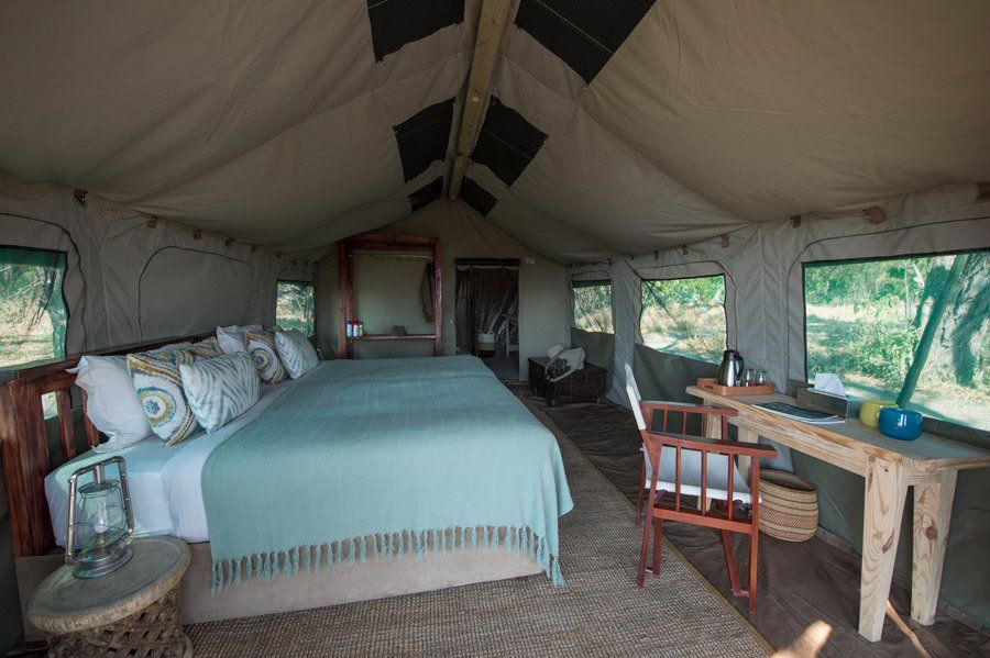 Safari tent interior with a large bed, desk, and windows overlooking greenery.