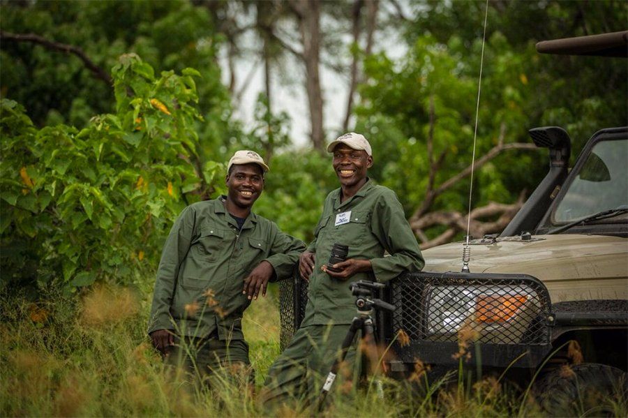 Two men in green uniforms smile next to a vehicle in tall grass and trees.