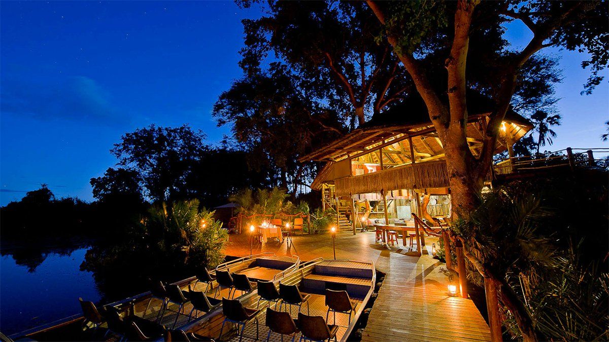 Wooden structure lit at night near water, with seating area and trees.