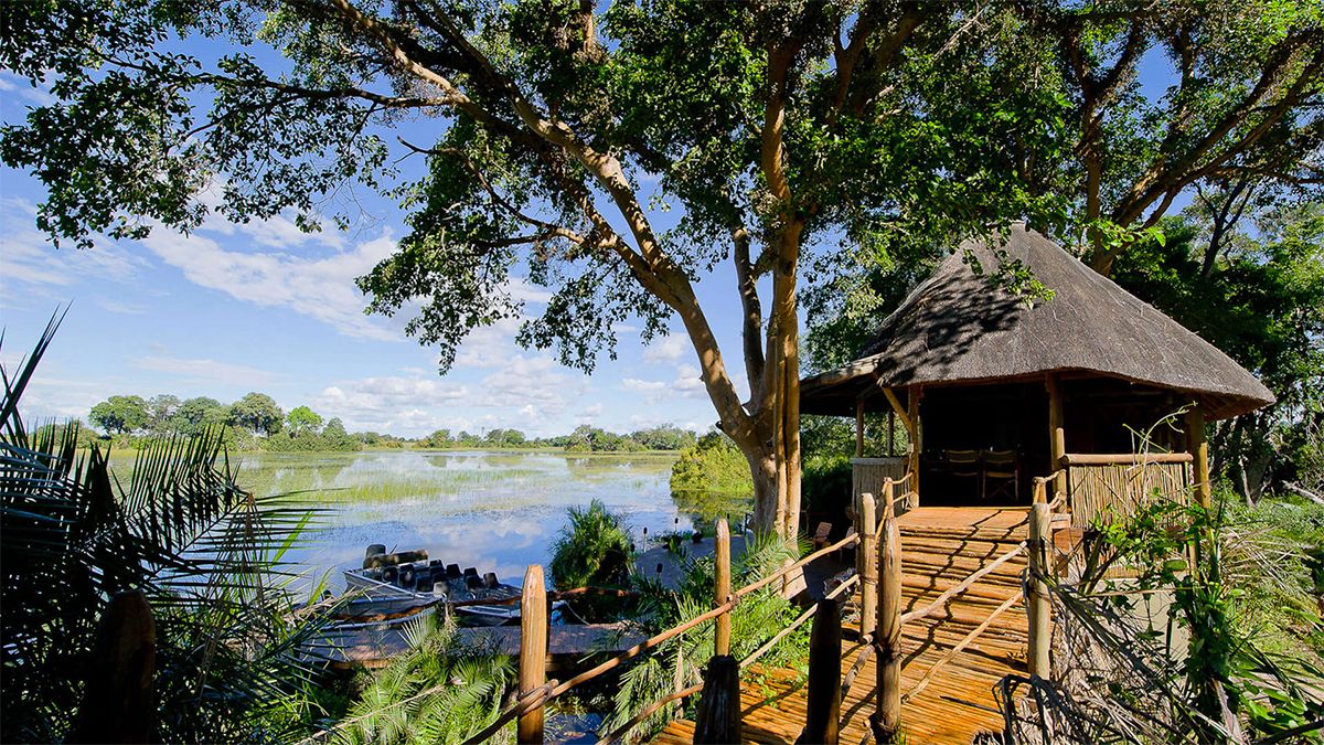 Wooden cabin with thatched roof overlooking a lake, surrounded by trees and a blue sky.