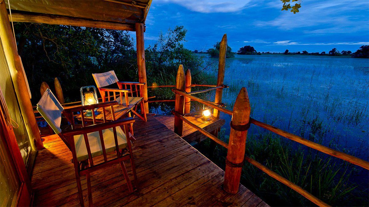 Wooden porch overlooking a calm body of water at dusk. Lantern-lit table with chairs.