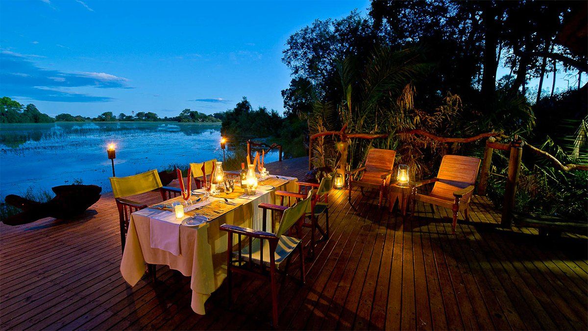 Outdoor dining setting, table set with candles overlooking water at dusk. Wooden deck, chairs.