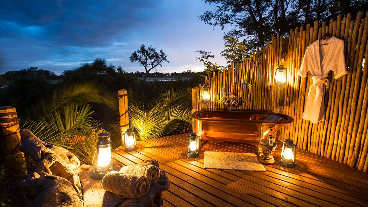 Outdoor copper bathtub with lit lanterns and robe against a reed fence at dusk.