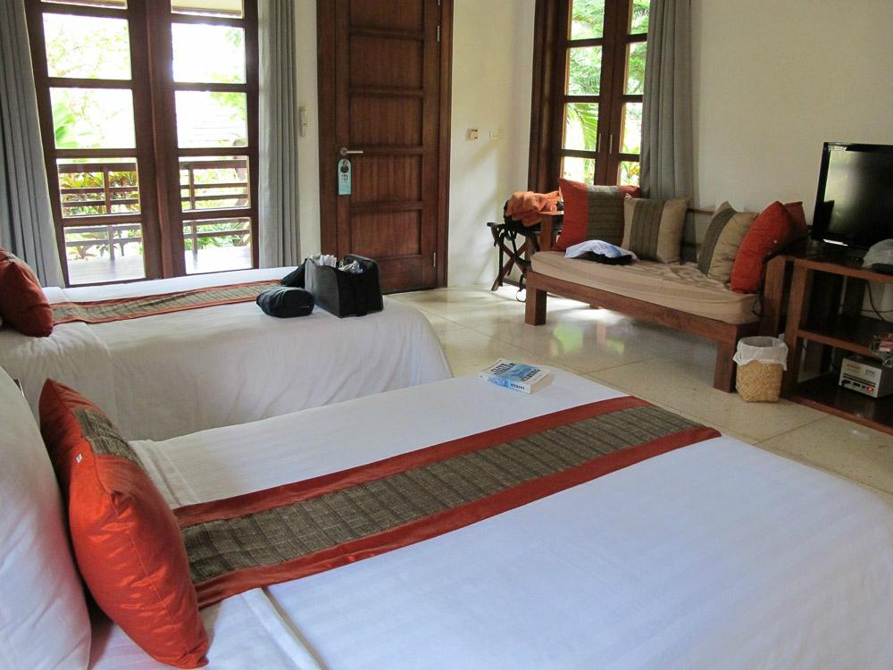 Two beds in a room with a couch, TV, and balcony door. White sheets, orange pillows, and wooden furniture.