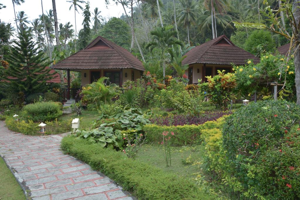 Two small buildings with brown roofs surrounded by lush greenery and a stone path.