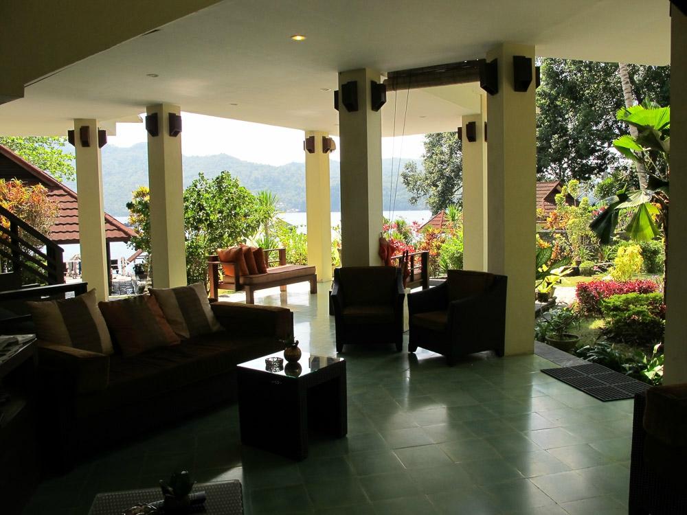 Covered outdoor seating area with sofas, armchairs, and a view of a lake and lush landscaping.