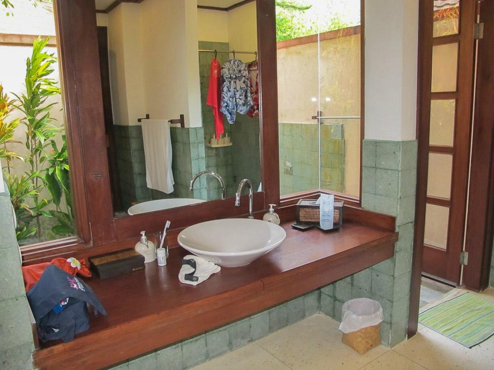 Bathroom with a large wooden counter, white sink, and open doorway to an outdoor shower area and foliage.