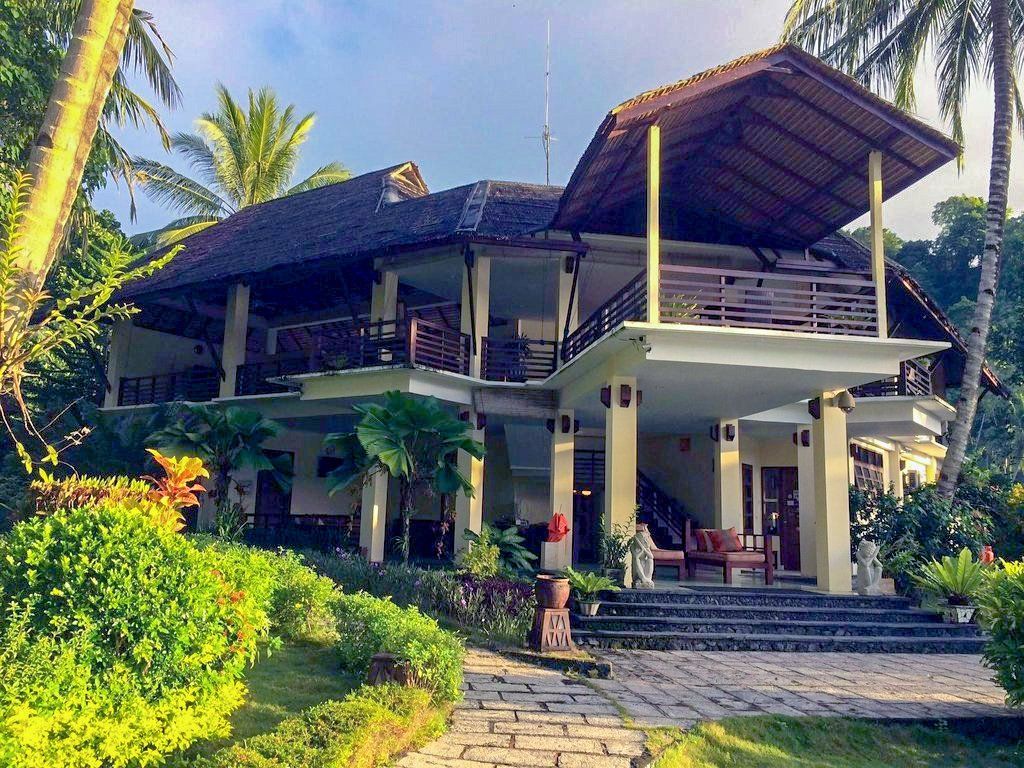 Tropical-style villa with thatched roof and balconies, surrounded by lush greenery and stone pathway.