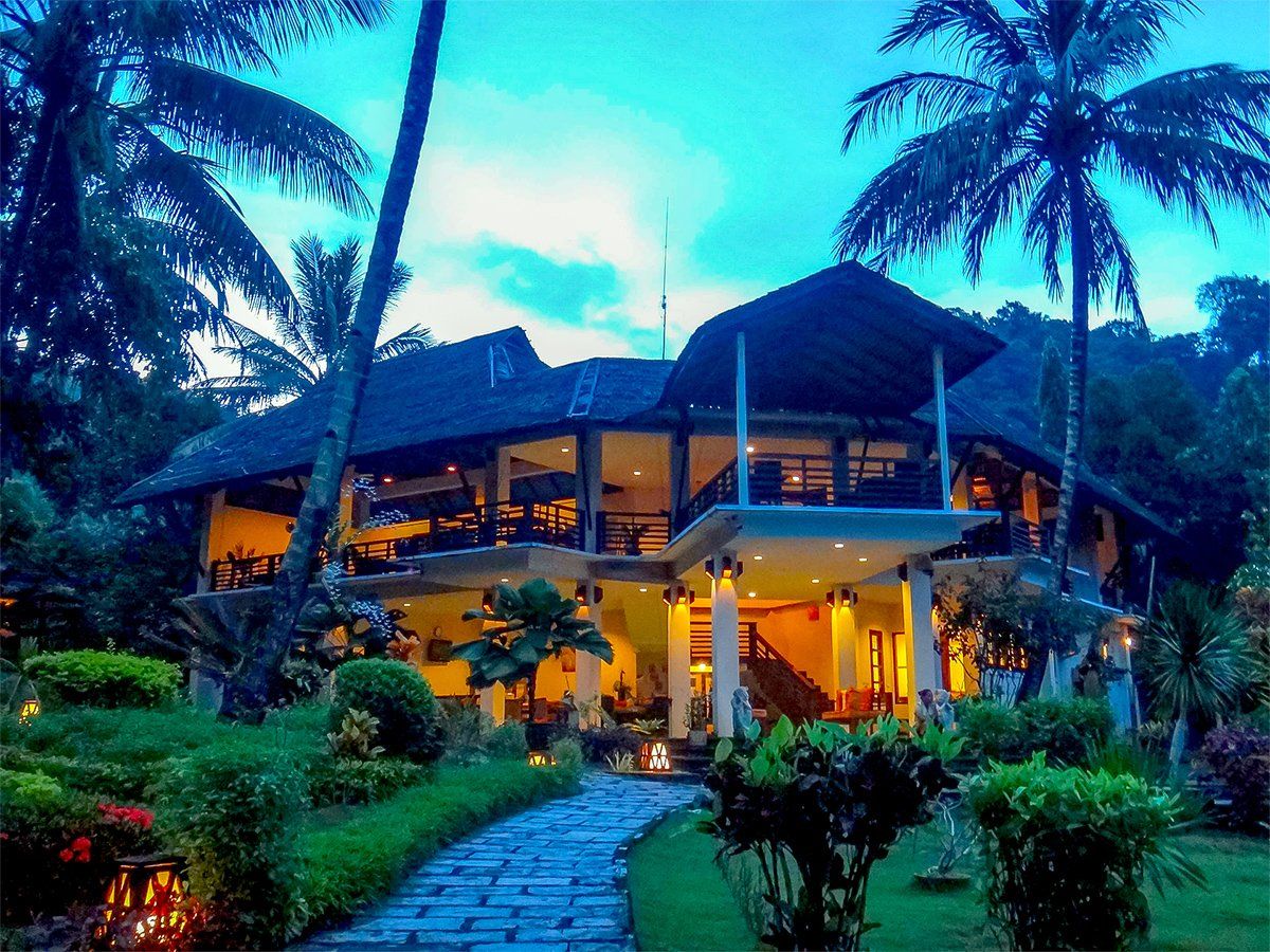 Tropical resort building with thatched roof, lit interior, palm trees and stone path leading to it.