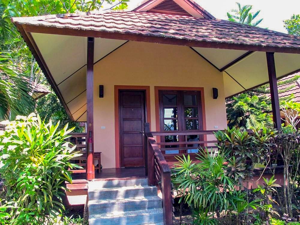Tan-colored bungalow with brown roof, porch, steps, and door, surrounded by lush green plants.