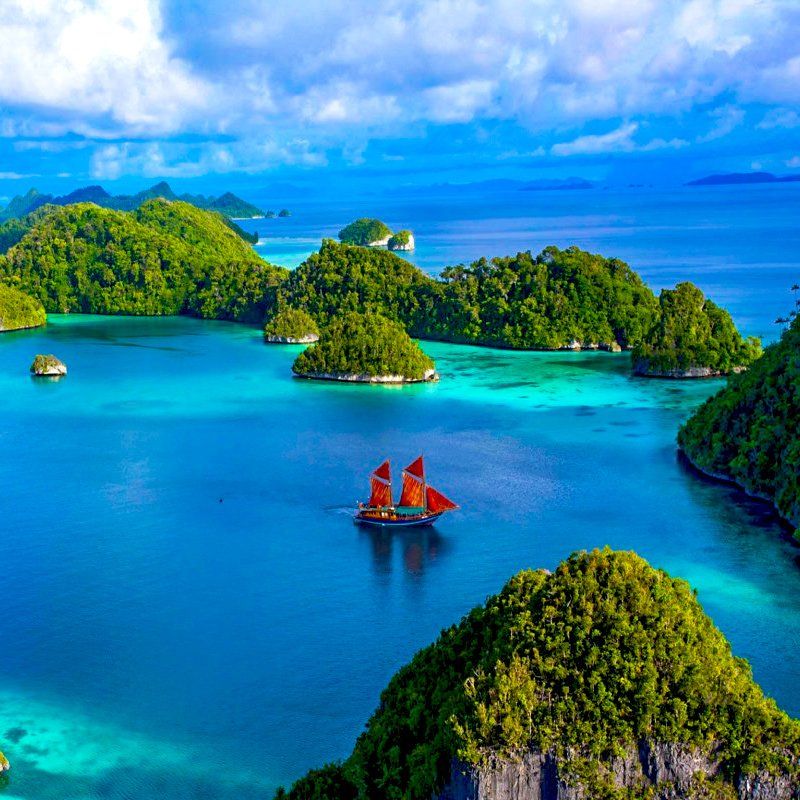 Emerald islands dot a turquoise sea; a red-sailed ship sails between them.