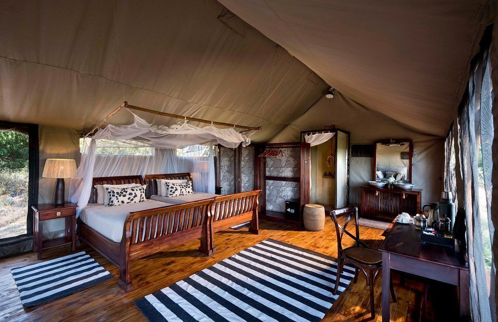 Safari tent interior with a bed, dresser, and desk. Striped rugs.