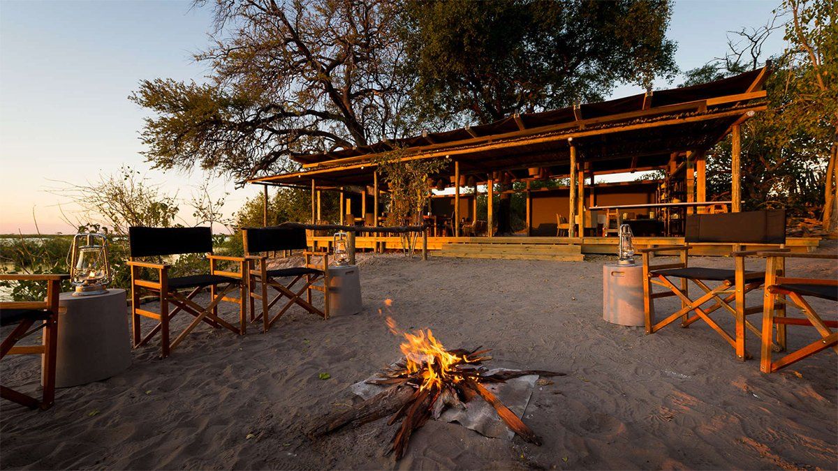 Campfire with chairs set in front of a covered porch overlooking a body of water, dusk setting.