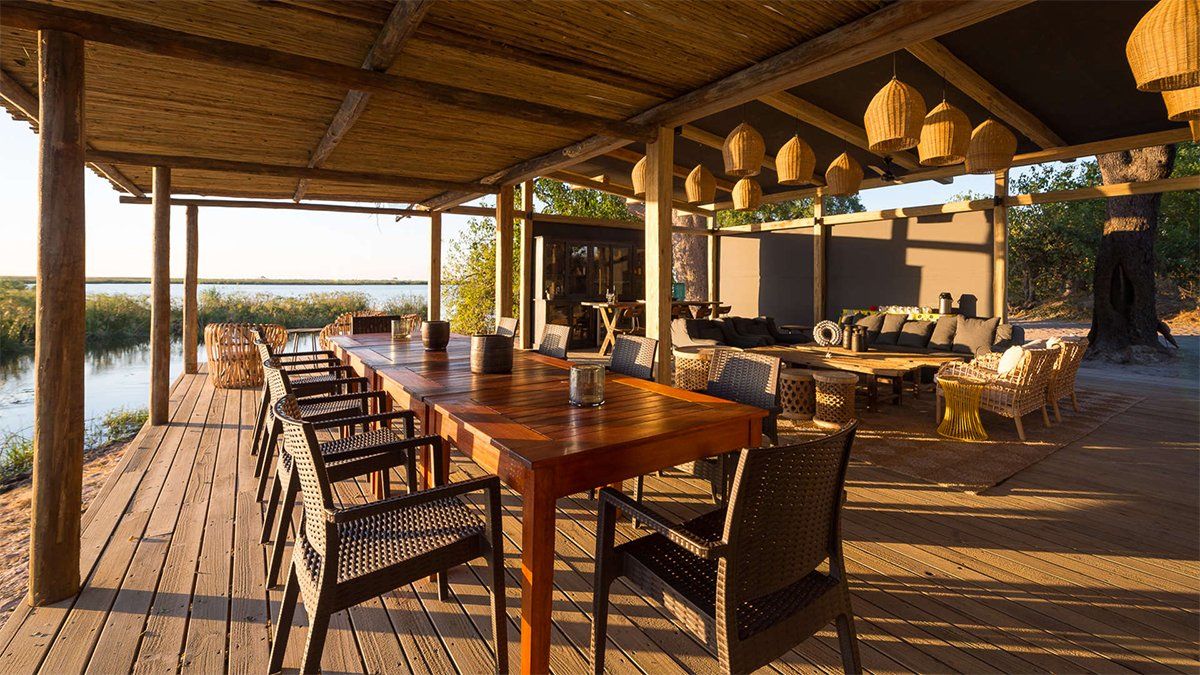 Outdoor dining area overlooking water, with a long wooden table, chairs, and woven roof.