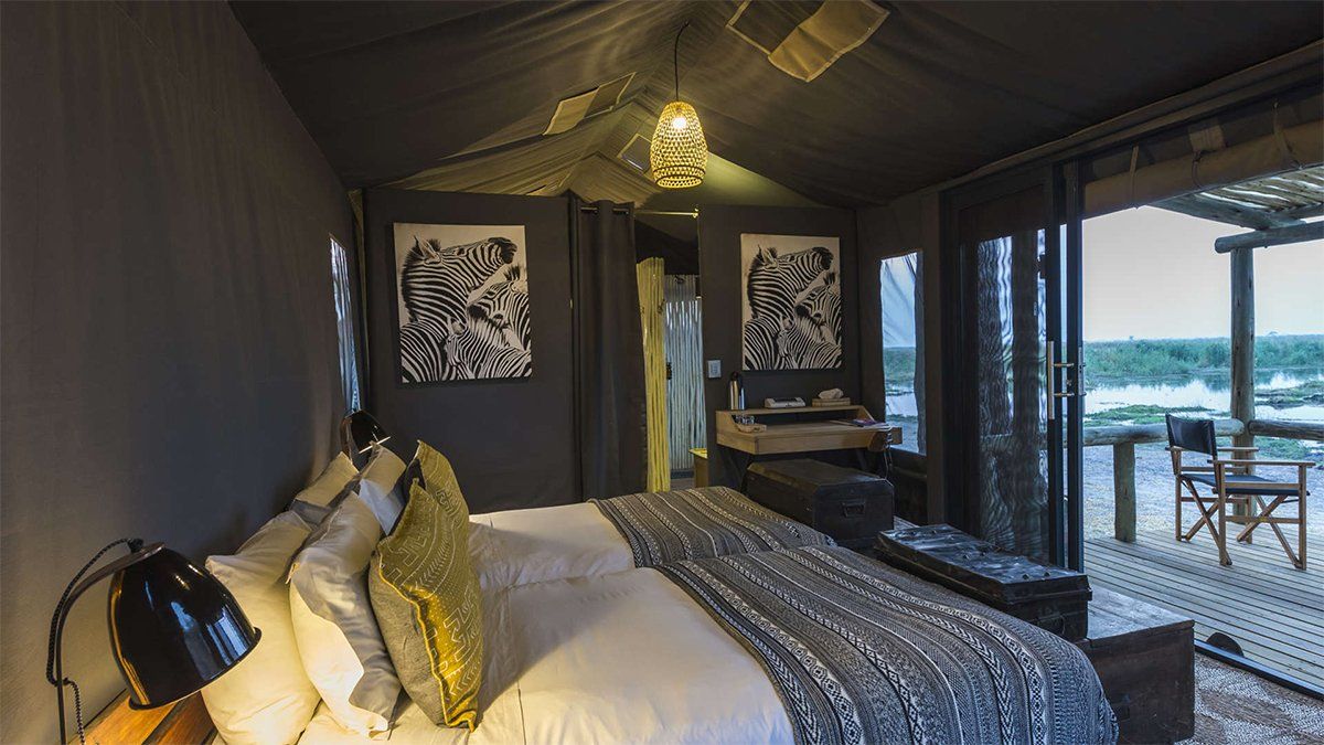 Bedroom in a tent, with a bed, art, desk, and view of a deck and water.