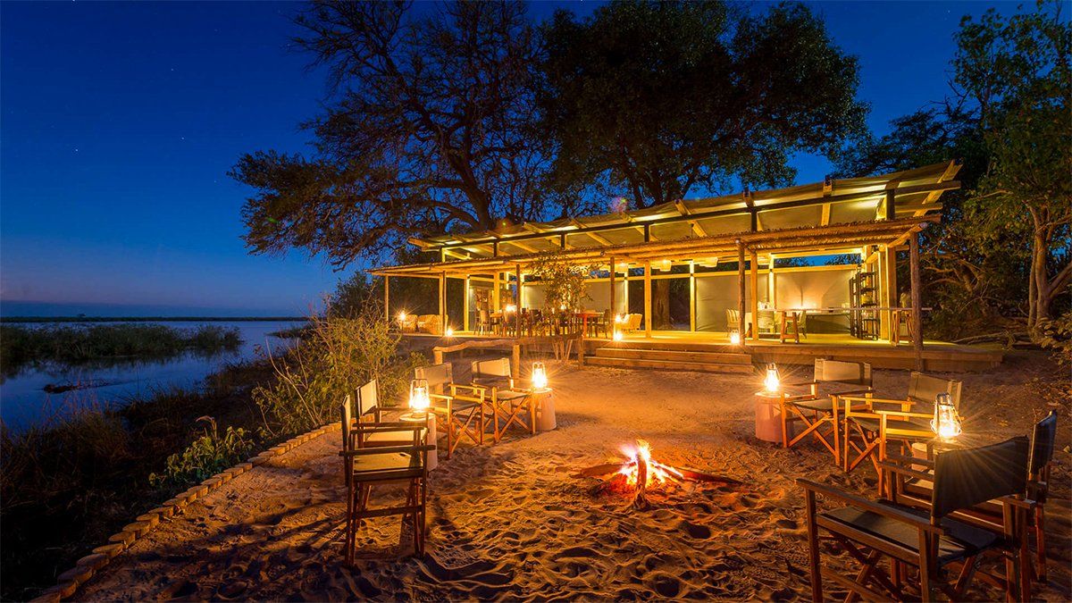 Lit-up lodge at dusk with bonfire and seating overlooking a calm body of water.