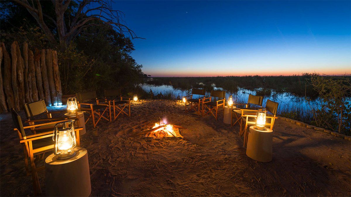 Campfire scene at dusk with chairs, lanterns, and a water view.